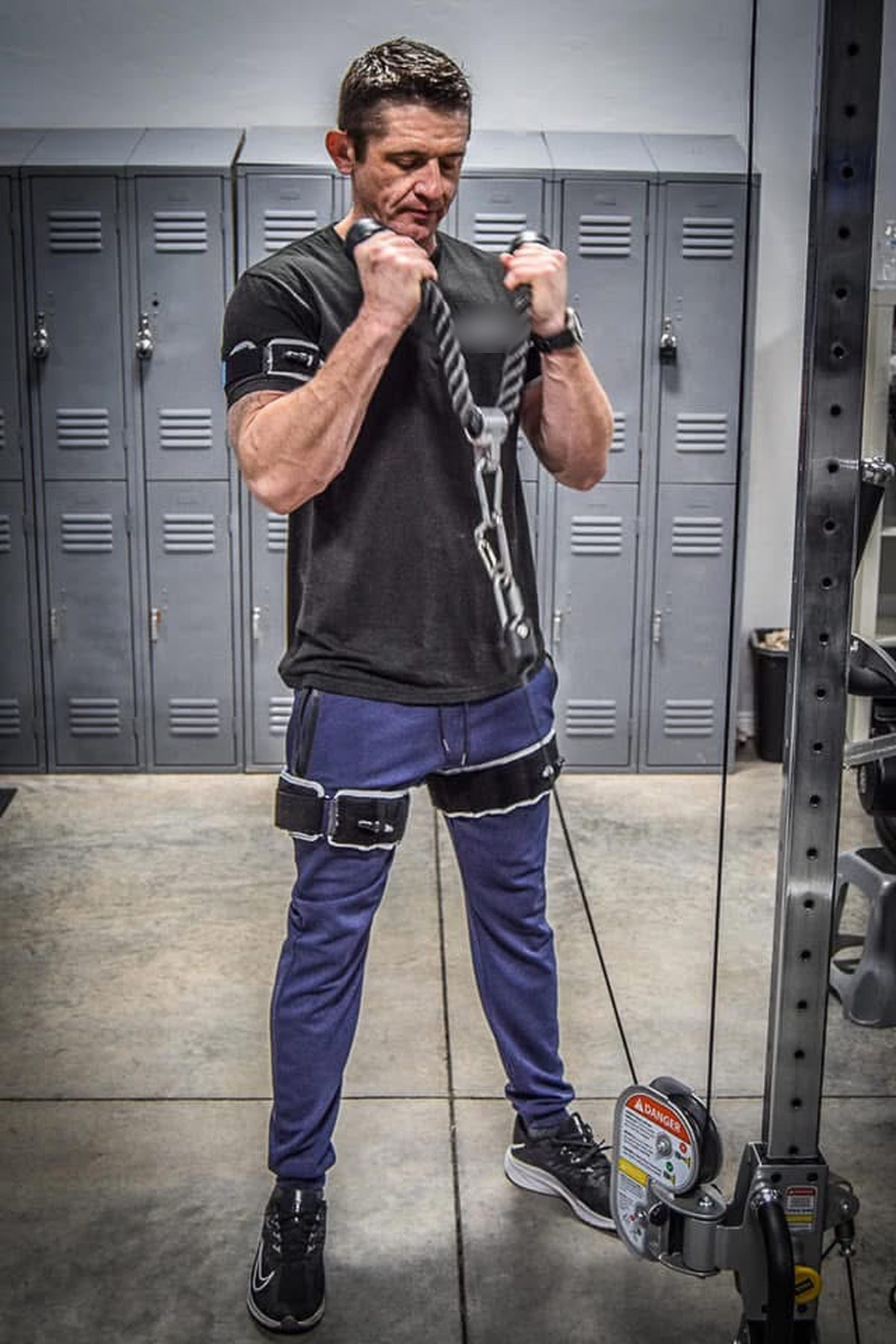 Ty McMillan doing rope-handle cable curls by gray lockers, wearing B3 BFR bands on his arms and legs