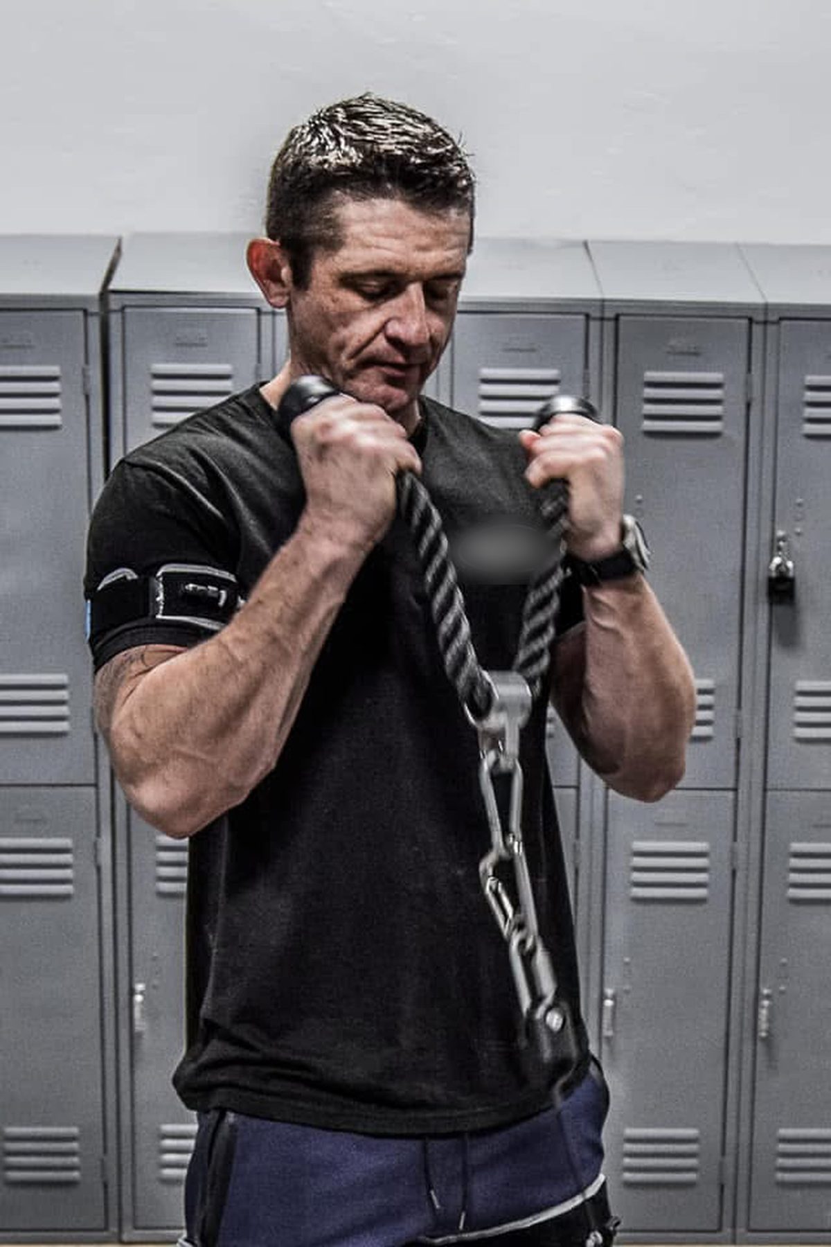 Ty McMillan doing rope-handle cable curls by gray lockers, wearing B3 BFR bands on his arms