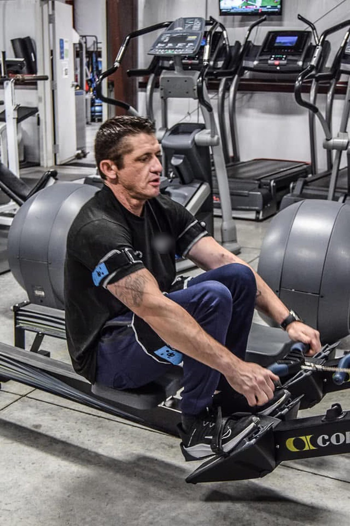 Ty McMillan on a rowing machine in a gym, wearing a B3 BFR band on his arm