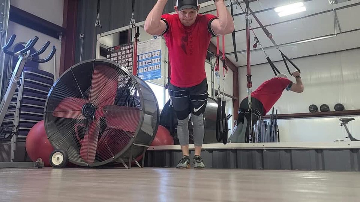 Ty McMillan in a red shirt on a suspension trainer in a gym, wearing B3 BFR bands on his arms and legs