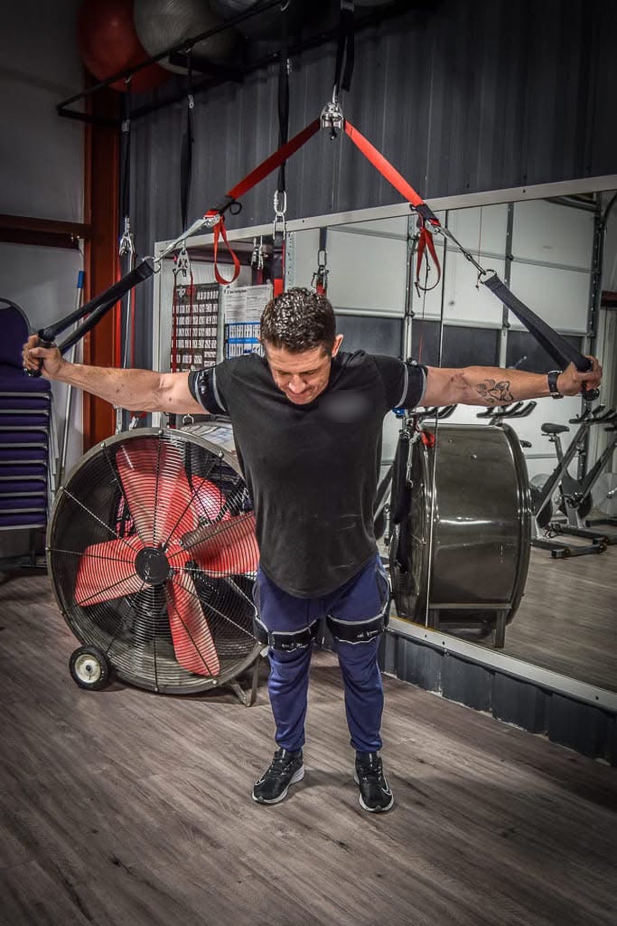 Ty McMillan doing a suspension-strap exercise in a gym, wearing B3 BFR bands on his arms and legs