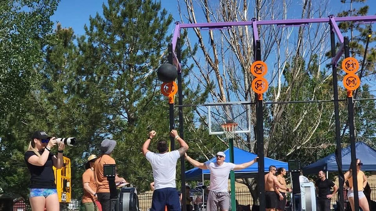 Ty McMillan, in a white shirt and navy shorts, throwing a medicine ball at a target on an outdoor rig while others watch